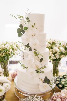 a wedding cake with white flowers and greenery on the top is surrounded by other floral arrangements