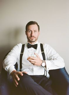 a man in a white shirt and bow tie sitting on a blue chair holding a drink