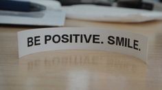 a white sticker that says be positive smile on top of a wooden table next to a computer keyboard