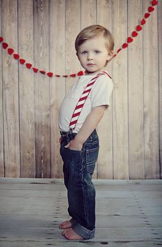 a young boy wearing a tie and jeans standing in front of a wooden wall with hearts on it