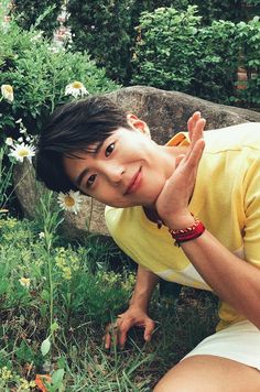 a young man sitting in the grass with his hand up to his face and looking at the camera