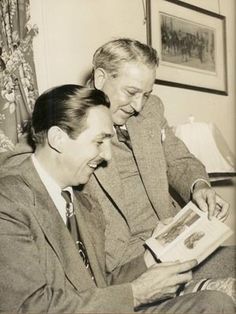 two men sitting next to each other looking at something in a book while one man holds his hand out