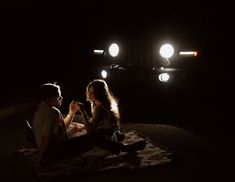 a man and woman sitting on the ground in front of a truck at night with their lights on