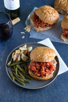 two sloppy joe sandwiches and green beans on a plate next to a bottle of wine