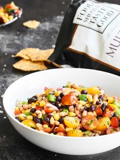 a white bowl filled with corn salad next to a bag of chips and a bottle of wine