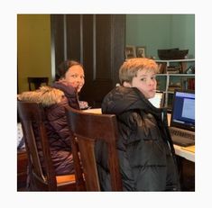 two people sitting at a table with a laptop computer in front of them and another person standing next to the desk