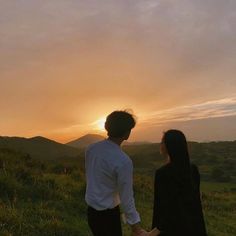 a man and woman holding hands while standing on top of a lush green hillside at sunset