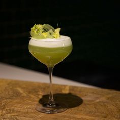 a green drink in a wine glass sitting on a wooden table with some flowers growing out of it