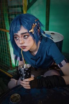 a woman with blue hair and piercings drinking from a black cup while sitting on the ground