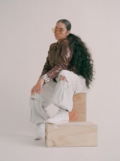 a woman sitting on top of a wooden block wearing white pants and a brown jacket