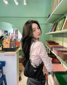 a woman standing in front of a bookshelf holding a black purse and looking at the camera