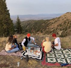 four people sitting on a blanket at the top of a hill eating food and drinking