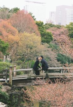 two people kissing on a bridge over a stream in a park with trees and buildings in the background