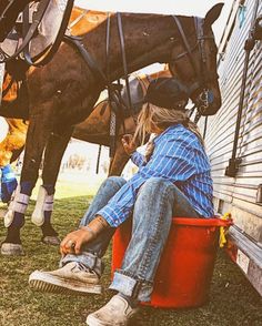 a woman sitting on the side of a house next to a brown horse and red bucket