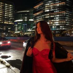 a woman in a red dress standing on the side of a road at night time