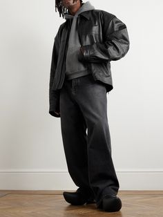 a man standing in front of a white wall wearing a leather jacket and black pants