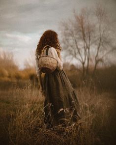 a woman standing in tall grass with a basket on her shoulder and back to the camera