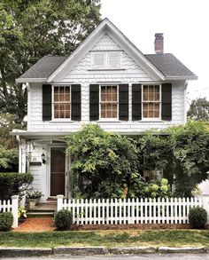 a white house with black shutters on the front and side windows, surrounded by greenery