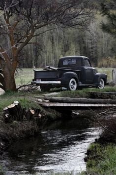 an old black truck parked on the side of a road next to a tree and bridge