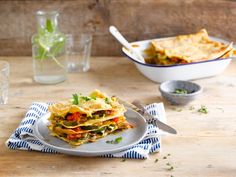 a white plate topped with layered lasagna next to a bowl of vegetables and a glass of water