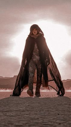 a woman walking across a sandy beach under a cloudy sky with her arms wrapped in a black shawl