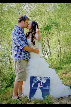 a bride and groom kissing in front of a photo album on the grass with trees behind them