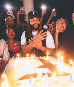 a group of people standing around a birthday cake with candles on it and one person holding his hands up