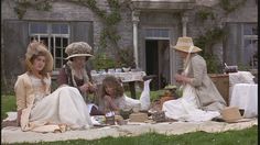 three women dressed in period clothing sitting on the grass outside an old house with picnic tables and chairs