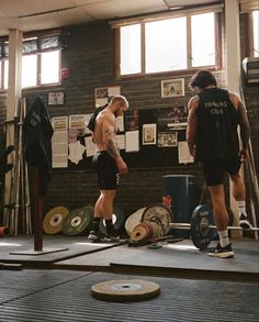 two men standing in a gym with one holding a barbell and the other looking down