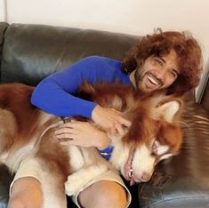 a man sitting on top of a couch next to a brown and white husky dog