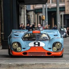 an orange and blue race car is parked on the side of the street while people walk by