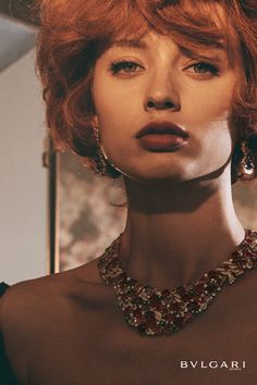 a woman with bright red hair wearing a necklace and earrings on her neck is looking at the camera