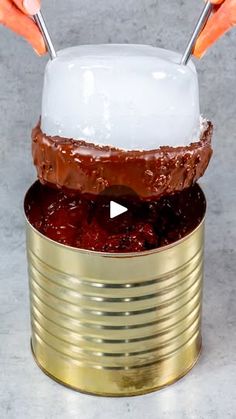 a person holding two spoons over a chocolate cake in a gold tin with white frosting