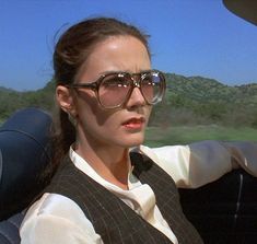 a woman driving in the back seat of a car wearing sunglasses and a vest, with mountains in the background