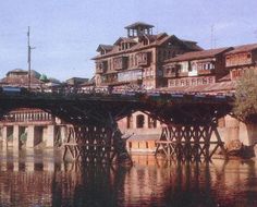 an old wooden bridge over a body of water with buildings in the background and people walking on it