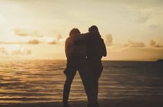 two people standing next to each other in front of the ocean at sunset or sunrise