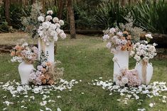 three white vases with flowers are on the grass in front of some trees and bushes