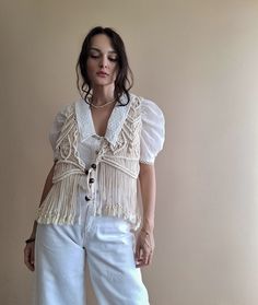 a woman standing in front of a wall wearing white pants and a top with tassels