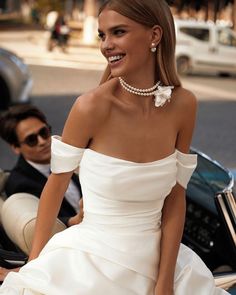 a woman in a white dress is sitting on a car and smiling at the camera