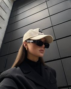 a woman wearing sunglasses and a hat stands in front of a wall with black tiles