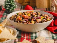 a white bowl filled with beans and vegetables next to tortilla chips on a plaid table cloth