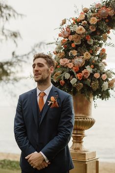 a man in a suit and orange tie standing next to a tall flower arrangement on a pedestal