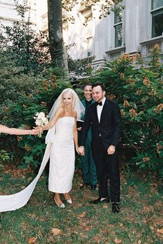 the bride and groom are holding hands with their friends in front of a building that is surrounded by greenery