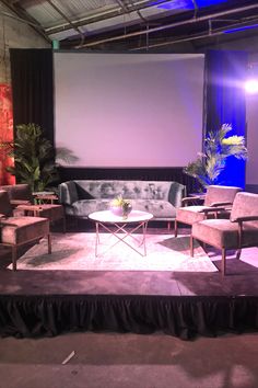 a stage set up with chairs, couches and a table in front of a projector screen