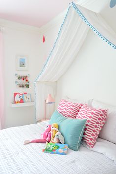 a white bed topped with lots of pillows next to a pink curtain and window sill