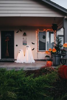 two white ghost statues in front of a house decorated for halloween with lights and pumpkins