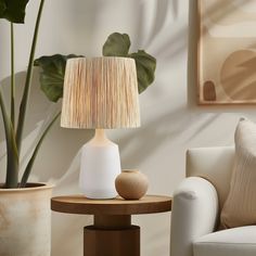 a table lamp sitting on top of a wooden table next to a white chair and potted plant