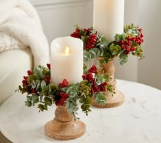 two white candles are sitting on a table with holly and red berries in vases