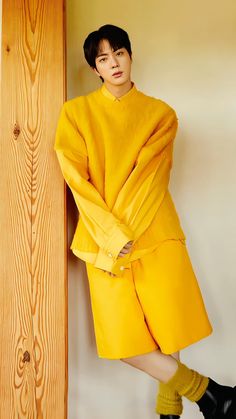 a woman leaning against a wooden wall wearing a yellow coat