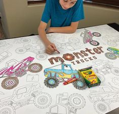 a young boy sitting at a table with monster trucks on it and coloring the paper